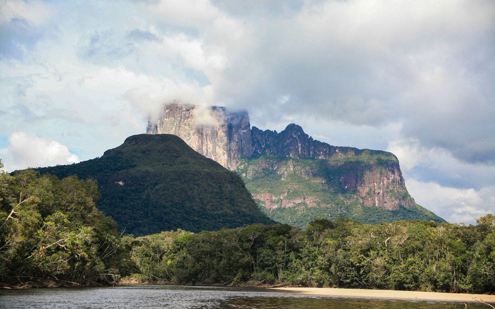 Angel Falls seen small against the full height of Auyán-tepui's sandstone wall, two adult figures standing on the valley floor in the foreground for scale, Canaima National Park